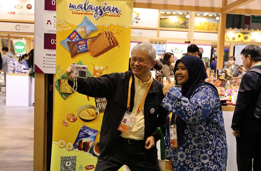 Um expositor de Cingapura (à esquerda) tira selfies com uma expositora da Malásia na área de exposição de produtos alimentares e agrícolas durante a 8ª Exposição Internacional de Importação da China (CIIE), em Shanghai, leste da China, em 7 de novembro de 2025. (Xinhua/Fang Zhe)